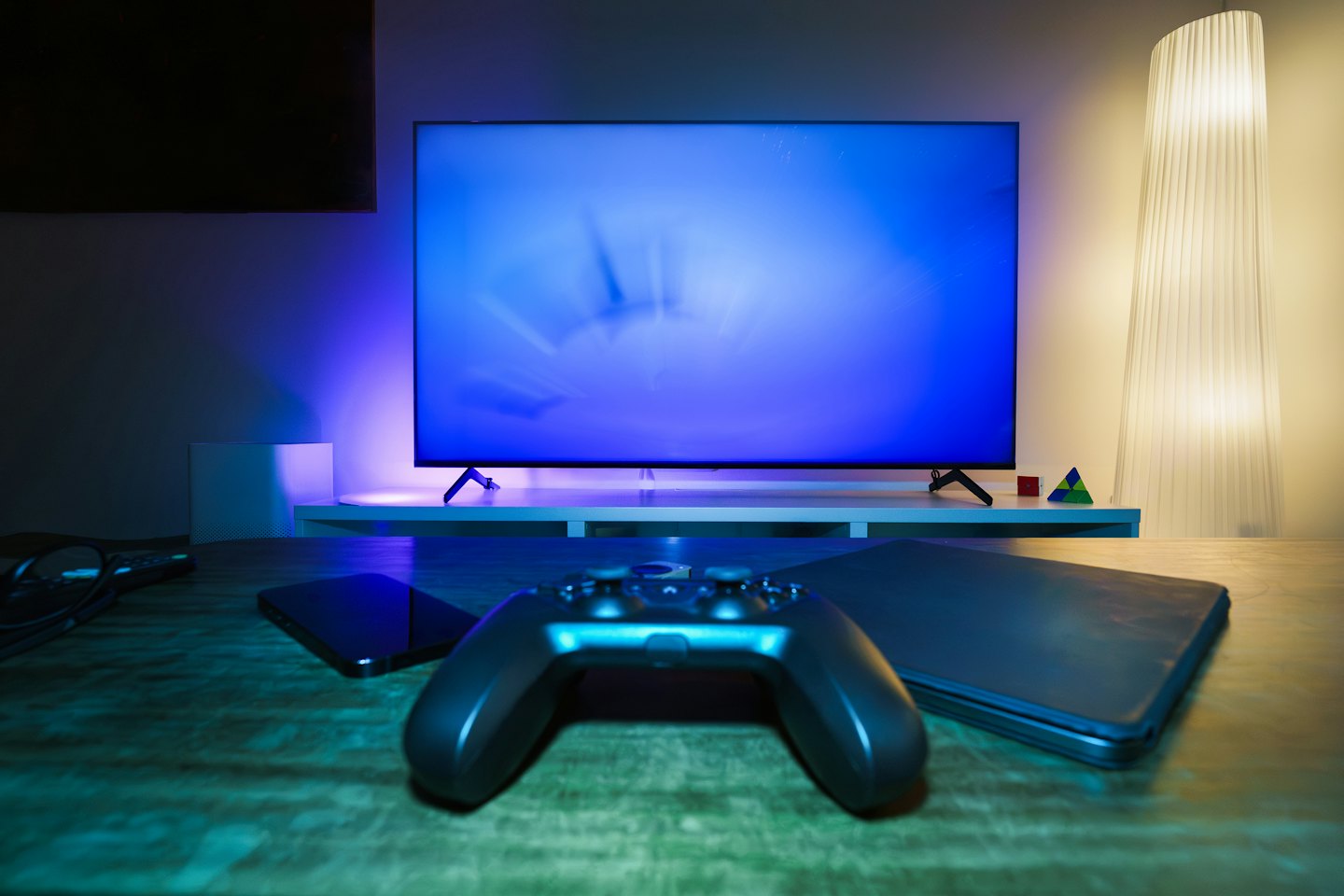 Game controller, phone, and laptop on a table with a glowing TV.